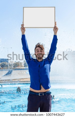 Bearded attractive inviting young man holding a framed blank whiteboard in blue sky