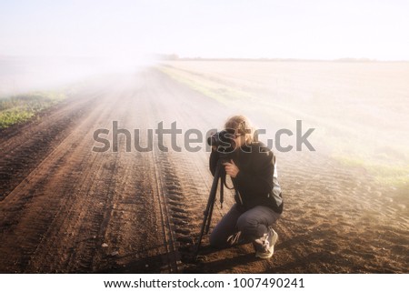 A woman in her mid fifties in casual clothes kneeling down with her camera and a tripod taking pictures of a fire with drifting smoke in a rural agricultural autumn landscape