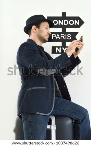 Traveler man in front of a sign with different cities using his mobile phone
