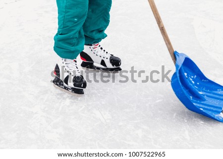 sportsman on skates cleans a blue plastic shovel on a wooden ice handle on a natural ice rink outdoors.
