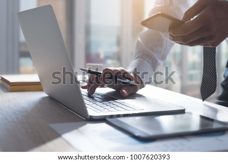 Business man using  mobile smart phone, busy working on laptop computer browsing internet or checking internet application on smartphone with digital tablet and business data on office desk, close up