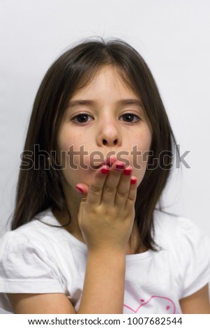 Cute little girl with hand on mouth blows a kiss to camera over white background. Pink polish nails. Charming love gesture