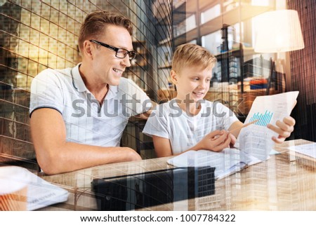 Lets compare. Cheerful kid expressing positivity while demonstrating his project and sitting near his father