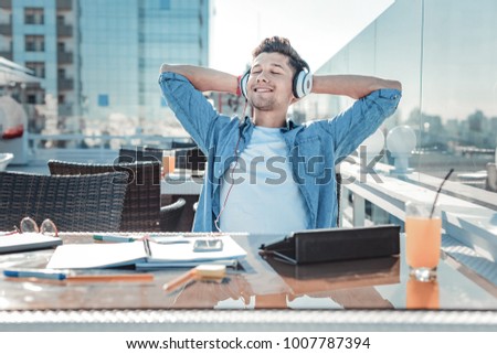What a wonderful day. Relaxed young man smiling and closing his eyes while listening to music playing in his headphones after studying hard. Royalty-Free Stock Photo #1007787394