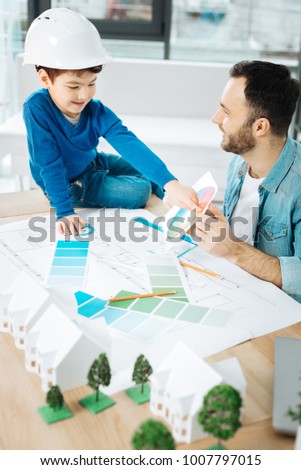 Crucial advice. Charming young architect sitting next to his little son and discussing the color choice with him while the boy pointing at color in the color chart Royalty-Free Stock Photo #1007797015