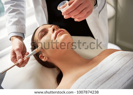 Unrecognizable male beautician applying facial mask on female face with eyes closed during cosmetic procedure.