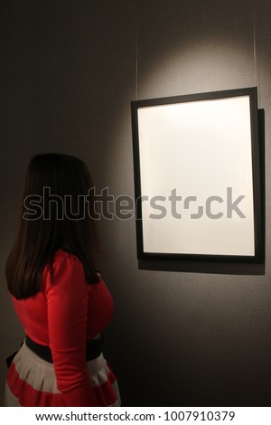 One blank white canvas frame to hang on the wall with the light shining down. The dark silhouette of a girl in a red dress looking at a picture or photo.