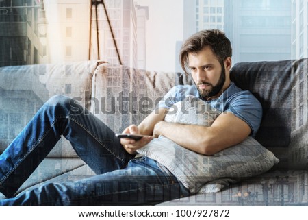 Lonely day. Disappointed unshaken depressed man lying on the sofa hugging the pillow and holding the control desk. Royalty-Free Stock Photo #1007927872