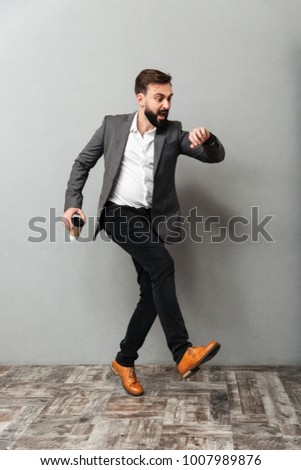 Full-length image of uptight man with takeaway coffee looking at watch being late and running along gray background Royalty-Free Stock Photo #1007989876
