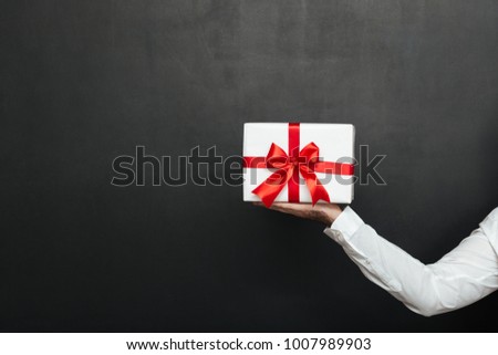 Cropped image of male hand holding white present box with red bow isolated over dark gray wall
