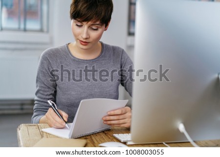 Busy young business woman writing at office desk with computer screen in foreground