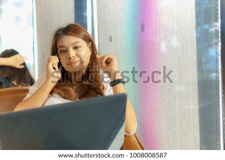 beautiful asian young business woman sitting near transparent window and using laptop for her job while talking with customer on the phone at cafe or coffee shop. happy or success business concept