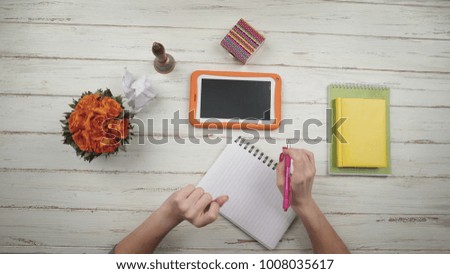 A girl writes a letter to a friend on a white wooden table next to flowers and orange digital tablet. Top view. Hands close up view