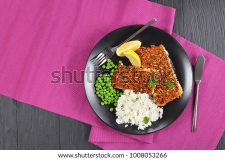 close-up of delicious white fish fillet  baked with crushed sunflower and pumpkin seeds spicy top crust served with rice and green peas on black plate with fork and knife, view from above