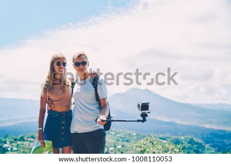 Young hipster guy holding monopod with camera making selfie standing with girlfriend traveling together, stylish tourists couple enjoying active leisure recreating on journey on island taking picture