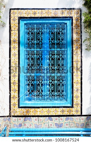 An ancient window with an ornament and a blue iron grating in Arabic style against a white wall.