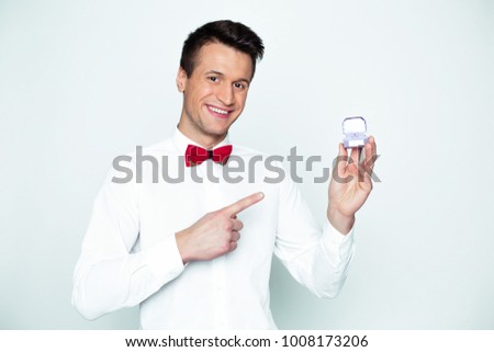 The most beautiful engagement ring! Beautiful smiling modern elegant man in a white shirt and red bow tie points a finger at a wedding a box in his hand.