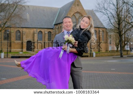 Couple, love, man in suit and blond woman with a wedding bouquet in black fur winter coat outside, Holland, dutch street and old catholic church on background