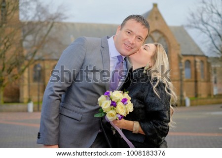 Couple, love, man in suit and blond woman with a wedding bouquet in black fur winter coat outside, Holland, dutch street and old catholic church on background