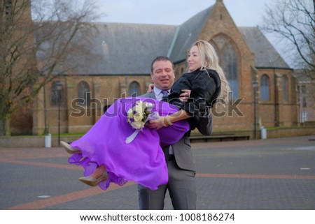Couple, love, man holding in arms blond woman with a wedding bouquet in black fur winter coat and long violet dress outside, Holland, dutch street and old catholic church on background