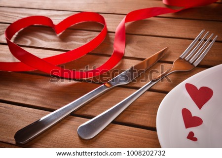 Table decorated with red ornaments for valentines day with hearts on a plate, cutlery and red ribbon forming a heart on planked wood table background. Elevated view