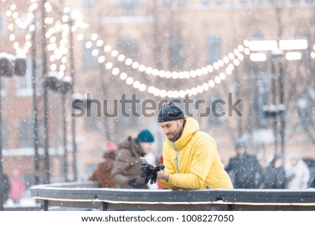 Portrait of joyful bearded man in yellow jacket, black hat standing near fence on ice rink and using smart phone, outdoors at snowy day/Winter time concept/ Weekend activities outdoor in cold weather