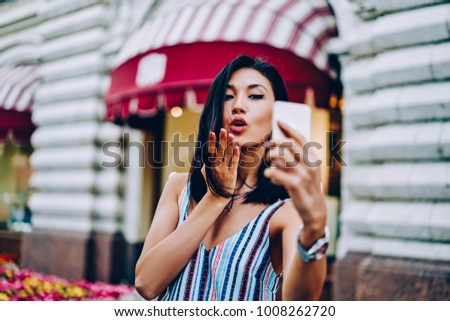 Attractive asian woman posing for selfie making air kiss standing on urban setting background, beautiful international tourist sending lips picture to boyfriend taking image using mobile application