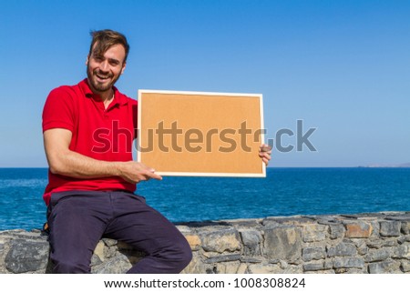 Bearded attractive inviting young man holding a framed blank corkboard in blue sky