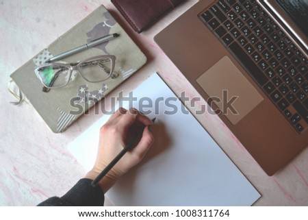 Office table with laptpo, notebook, pen, pencil, glasses, wallet and white paper. Stylish office tems. Woman writing on the white paper.