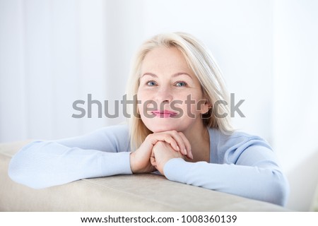 Active beautiful middle-aged woman smiling friendly and looking in camera in living room. Woman's face closeup. Realistic images without retouching with their own imperfections. Selective focus. Royalty-Free Stock Photo #1008360139