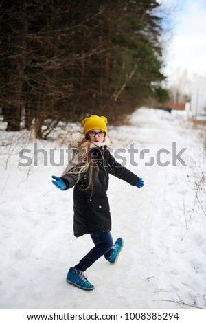Stylish pre teen girl 10-12 year old wearing yellow knitted hat and bblack jacket and glasses posing in forest outdoors. Having fun in winter forest