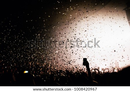 Confetti falling in front of the stage. Silhouettes of hands in front of highly illuminated stage. Concert atmosphere.
