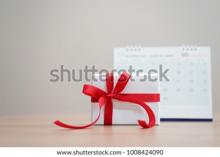 close up on white gift box with red ribbon and bow on wood table desk with calendar for special day concept