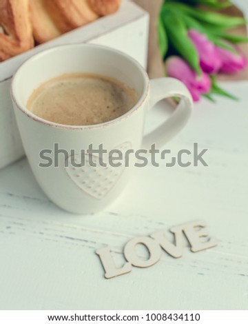 Cup of coffee with croissants, bouquet of pink tulips and wooden word LOVE on white shabby chic table. Romantic Valentines morning concept. Valentine's day breakfast.