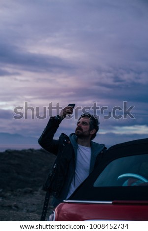 Handsome hipster man with beard stands next to cabrio car and makes photos on smartphone of beautiful and inspiring mesmerizing sunset skies with purple and pink color palette