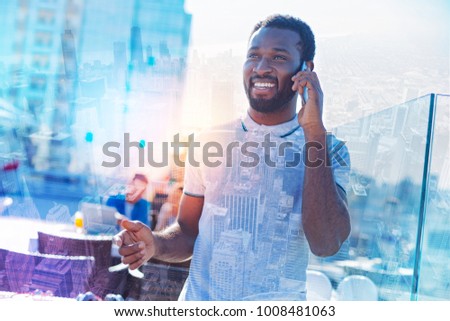 On cloud nine. Good-looking exuberant well-built afro-american man smiling and talking on his phone while standing near the table