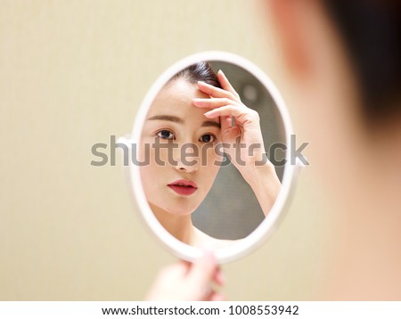 beautiful young asian woman looking at self in mirror, hand on forehead. Royalty-Free Stock Photo #1008553942