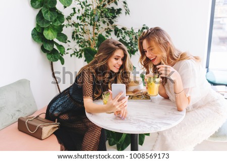 Two long-haired girls resting in cafe with modern interior and laughing. Indoor portrait of funny smiling ladies in trendy clothes making selfie while drink fruit cocktails. 