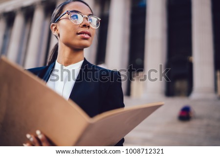 Thoughtful African American businesswoman in optical spectacles holding documents looking away while standing in urban setting with copy space. Female dark skinned student of high economic university Royalty-Free Stock Photo #1008712321