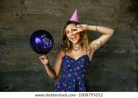 Winking beautiful funny celebrating birthday, showing peace gesture, holds a balloon. Wearing dress, festive hat. Holiday concept.