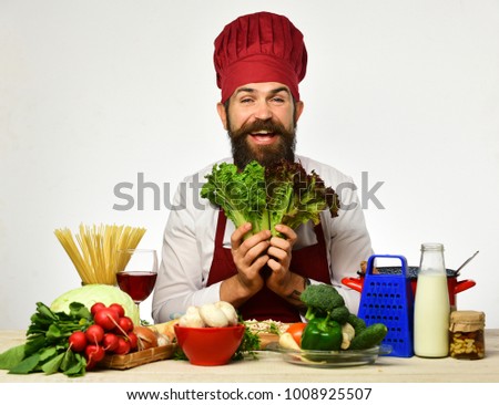 Chef prepares meal. Cook with cheerful face in burgundy uniform sits by kitchen table with vegetables and kitchenware. Man with beard holds lettuce on white background. Restaurant cuisine concept.