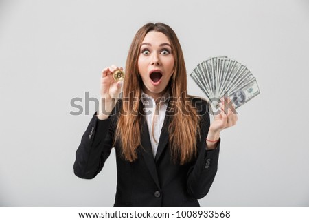 Shocked beautiful businesslady in official suit holding golden bitcoin and dollars in hands and looking camera isolated