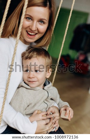 Charming mother has fun with her son playing on the swing in cosy room