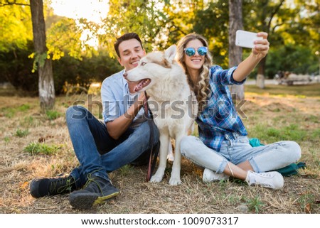 young stylish couple playing with dog in park, man and woman happy family together, husky breed, summer style, sunny, positive romantic mood, making selfie photo on phone