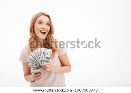 Picture of excited young lady looking aside isolated over white background holding money using mobile phone.