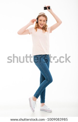 Photo of cheerful young lady standing isolated over white background. Looking aside listening music using mobile phone.
