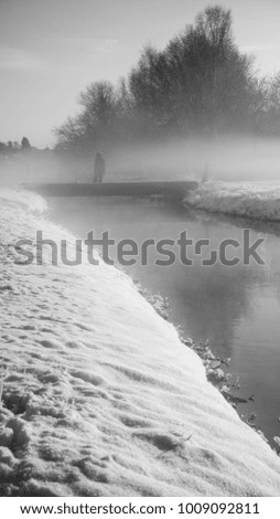 Black and White Picture of a river and a person standing on a bridge with fog covering it.