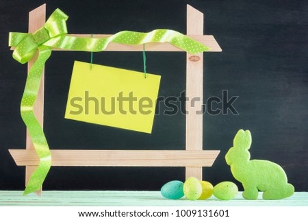 Easter card with painted eggs, a bunny shaped cookie and a wooden frame with ribbon and bow and a blank green message card, on a black background.