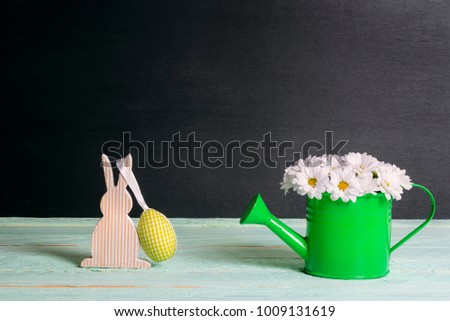 Cute white daisies arrangement in a watering can and a wooden bunny with a yellow fabric egg hanging, on a green table and a black background.