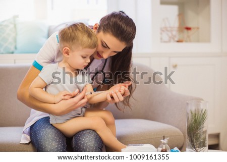 The young nurse sitting on the sofa in the room and holding the cute little boy on the lap and she showing him as give the shot
